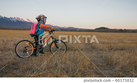 One caucasian children walk with bike in wheat field. Little girl walking black orange cycle on background of beautiful snowy mountains. Biker stand with backpack and helmet. One caucasian children walk with bike in wheat field. Little girl walking black orange cycle on background of beautiful snowy mountains. Biker stand with backpack and helmet. 60173832