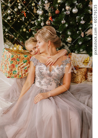 Daughter hugging her mother while sitting at christmas tree with gift boxes 60179135