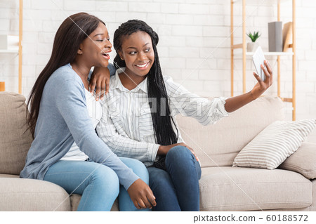 Cheerful black girls taking selfie while sitting on couch at home 60188572