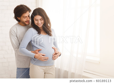 Loving man hugging his pregnant wife from behind standing near window 60189113