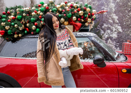 Beautiful young girl is wearing winter clothes holding sparklers near red car with decorated xmas tree on the roof, holiday and new years concept. Beautiful young girl is wearing winter clothes holding sparklers near red car with decorated xmas tree on the roof, holiday and new years concept. 60190952