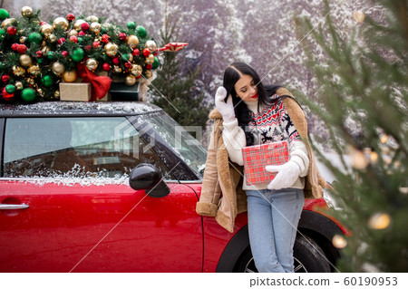 Beautiful young girl is wearing winter clothes holding gift box near red car with decorated xmas tree on the roof, holiday and new years concept. 60190953