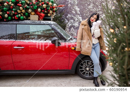 Beautiful young girl is wearing winter clothes near red car with decorated xmas tree on the roof, holiday and new years concept. 60190954
