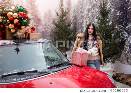 Beautiful young girl is wearing winter clothes holding gift box near red car with decorated xmas tree on the roof, holiday and new years concept. 60190955