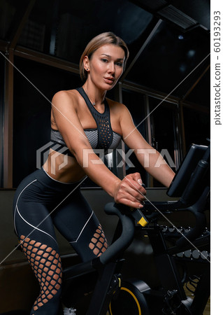 Young confident woman athlete exercising on bicycle indoors. Attractive determined fitness girl doing cycling exercises in dark gym. Functional workout training of sports girl. Cardio training 60193293