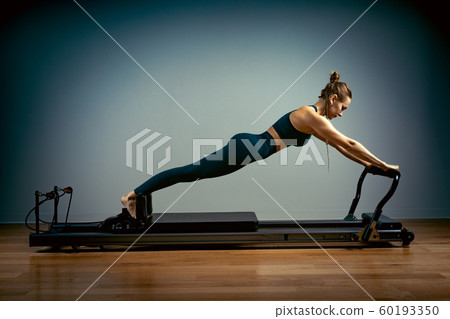 Young girl doing pilates exercises with a reformer bed. Beautiful slim fitness trainer on reformer gray background, low key, art light. Fitness concept 60193350