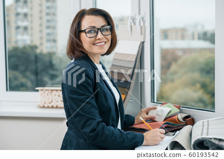 Female decorator interior designer at workplace with fabric samples. Smiling female in jacket, glasses looking at the camera standing near the window Female decorator interior designer at workplace with fabric samples. Smiling female in jacket, glasses looking at the camera standing near the window 60193542