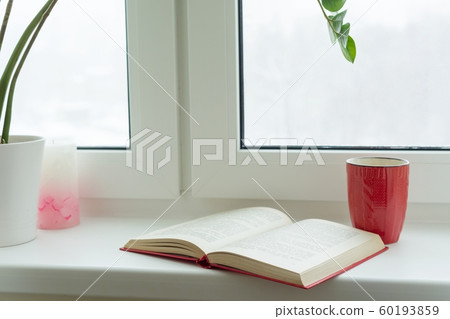 Winter time, snow-covered window, an open book and a cup with a hot drink on the windowsill 60193859
