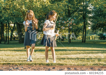 Two little girl friends schoolgirl 7, 8 years old play with soap bubbles on the meadow in the park. Two little girl friends schoolgirl 7, 8 years old play with soap bubbles on the meadow in the park. 60194235