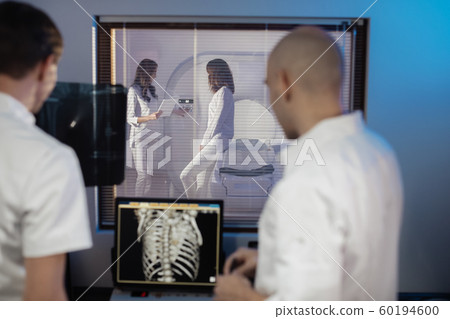 In Control Room Doctor and Radiologist Discuss Diagnosis while Watching Procedure, In the Background Patient Undergoes MRI or CT Scan Procedure. In Control Room Doctor and Radiologist Discuss Diagnosis while Watching Procedure, In the Background Patient Undergoes MRI or CT Scan Procedure. 60194600