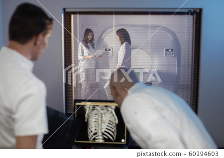 In Control Room Doctor and Radiologist Discuss Diagnosis while Watching Procedure, In the Background Patient Undergoes MRI or CT Scan Procedure. In Control Room Doctor and Radiologist Discuss Diagnosis while Watching Procedure, In the Background Patient Undergoes MRI or CT Scan Procedure. 60194603