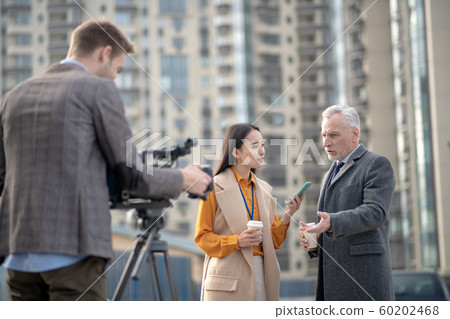 Female reporter in beige outfit asking questions to a grey-haired man 60202468