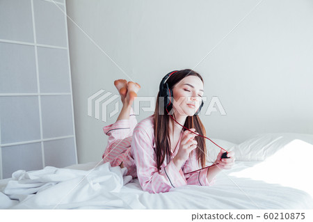 brunette in pink Pajamas listening to music with headphones on the bed brunette in pink Pajamas listening to music with headphones on the bed 60210875