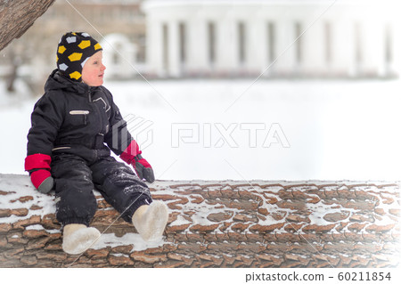 A little boy sits on a large fallen tree in winter A little boy sits on a large fallen tree in winter 60211854