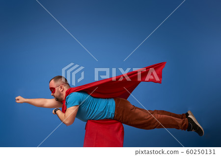 Male in red superhero cloak flying on empty blue background Male in red superhero cloak flying on empty blue background 60250131