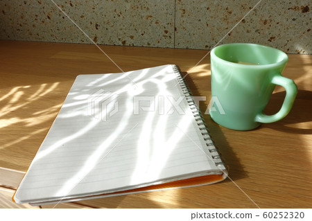 A cup and notebook with a cup of coffee and a blank notebook on the table shining through the curtain 60252320