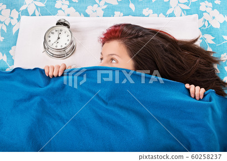 Girl hiding beneath a blanket looking at alarm clock Girl hiding beneath a blanket looking at alarm clock 60258237