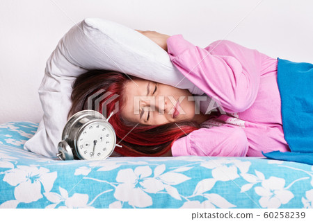 Annoyed girl covering her ears with a pillow from the alarm clock Annoyed girl covering her ears with a pillow from the alarm clock 60258239