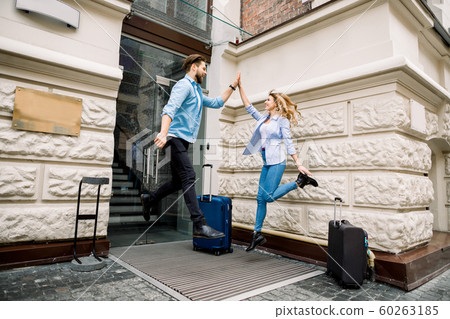 Young cheerful couple with suitcases jumping and giving five, having fun, before arriving to the hotel. Couple having fun outdoors, near the old city building Young cheerful couple with suitcases jumping and giving five, having fun, before arriving to the hotel. Couple having fun outdoors, near the old city building 60263185