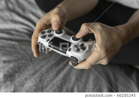 Gamer in white tshirt holding a gamepad in hands and sitting on a couch. Man hands playing a video game with a white controller. Play a video game at home with a pleasure. Gamer in white tshirt holding a gamepad in hands and sitting on a couch. Man hands playing a video game with a white controller. Play a video game at home with a pleasure. 60285245