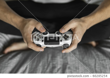 Gamer in white tshirt holding a gamepad in hands and sitting on a couch. Man hands playing a video game with a white controller. Play a video game at home with a pleasure. Gamer in white tshirt holding a gamepad in hands and sitting on a couch. Man hands playing a video game with a white controller. Play a video game at home with a pleasure. 60285254