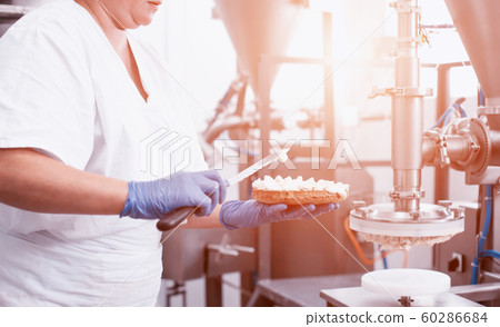 The pastry chef makes cake using modern equipment for the production of cakes. Line for spreading cream on biscuit cake. Industry The pastry chef makes cake using modern equipment for the production of cakes. Line for spreading cream on biscuit cake. Industry 60286684