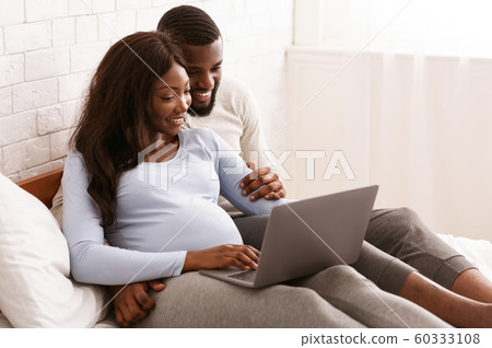 Pregnant woman and her husband using laptop, laying on bed Pregnant woman and her husband using laptop, laying on bed 60333108
