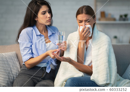 Girl Giving Pills And Water To Friend Sitting On Sofa Girl Giving Pills And Water To Friend Sitting On Sofa 60333252
