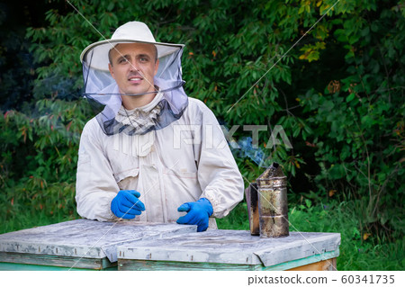 Young beekeeper working in the apiary. Professional beekeeper working outdoors and wearing the protective suits used for beekeeping. Beekeeper harvesting honey from a bee hive. 60341735