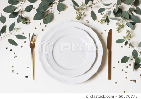 Christmas, New Year table place setting. Golden cutlery, porcelain plate, eucalyptus branches and golden confetti stars isolated on white background. Winter holidays background. Flat lay, top view. Christmas, New Year table place setting. Golden cutlery, porcelain plate, eucalyptus branches and golden confetti stars isolated on white background. Winter holidays background. Flat lay, top view. 60370775