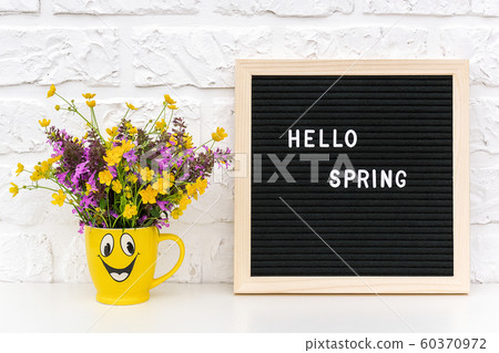 Text Hello Spring on black letter board and bouquet of colored flowers in funny yellow cup on background white brick wall. Concept Springtime mood and happiness 60370972