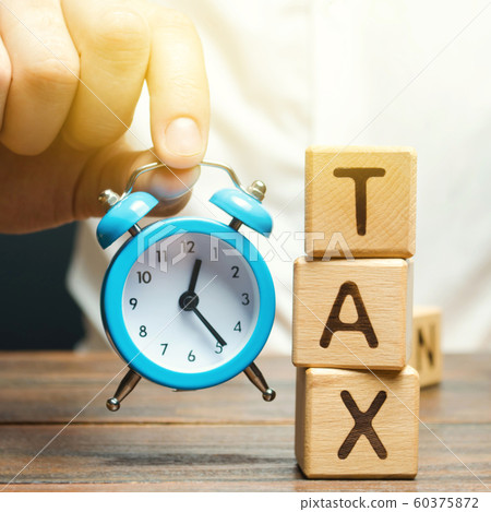 Businessman's hand holding a clock near the wooden blocks with the word Tax. Time to pay taxes. The concept of annual taxation. Taxes on vehicles, property, purchases, luxury. State fee Businessman's hand holding a clock near the wooden blocks with the word Tax. Time to pay taxes. The concept of annual taxation. Taxes on vehicles, property, purchases, luxury. State fee 60375872