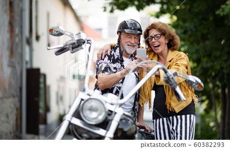 A cheerful senior couple travellers with motorbike in town, taking selfie. 60382293