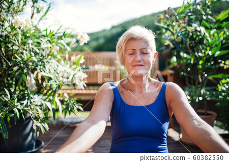 A senior woman sitting outdoors on a terrace in summer, doing yoga exercise. 60382550
