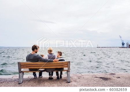 Rear view of young family with two small children on bench outdoors on beach. 60382659