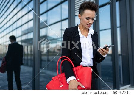 Caucasian woman in office clothes and with red bag waits colleague near office building Caucasian woman in office clothes and with red bag waits colleague near office building 60382713