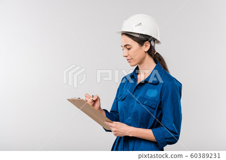 Young successful female engineer in blue workwear and safety helmet making notes Young successful female engineer in blue workwear and safety helmet making notes 60389231