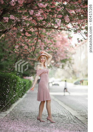 Young woman, dressed in white and yellow, in brimmed hat, stands among blossoming fruit trees and 60415166