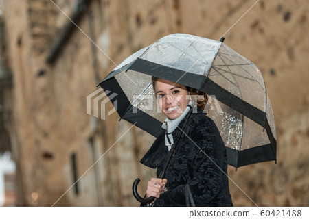 Dark-haired young woman walks through the streets of Caceres with a transparent umbrella on a rainy day Dark-haired young woman walks through the streets of Caceres with a transparent umbrella on a rainy day 60421488