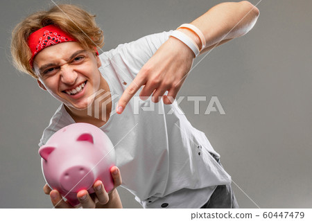 Young caucasian man in red sunglasses, white t-shirt with pink pig money box isolated on grey Young caucasian man in red sunglasses, white t-shirt with pink pig money box isolated on grey 60447479