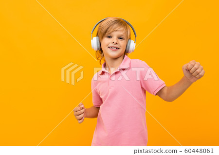 European blond boy in a pink T-shirt listens to music in white headphones on an orange background European blond boy in a pink T-shirt listens to music in white headphones on an orange background 60448061