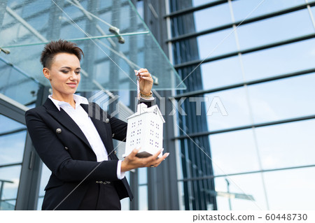 Caucasian woman in office clothes holds a little white house and waits colleague near the office 60448730