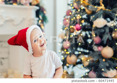 Adorable cute child boy having fun playing at home in red santa claus hat and festive christmas tree on background. Happy toddler kid looking ,smiling and dreaming for holiday gift, wonder an miracle 60471445