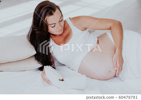a pregnant woman lies on a bed and reading a book 60478781