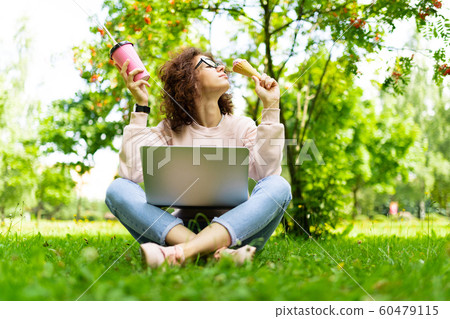 Pretty young caucasian woman is engaged in freelance in the park with the cup of coffe and ice-cream 60479115