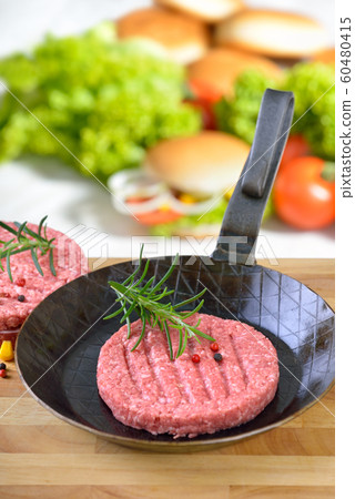 Raw burger patty in an iron frying pan ready to fry, some burger ingredients in the background 60480415