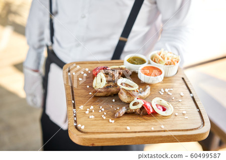 The waiter is holding a plate of Lamb tongue kebab with Pickled cabbage and two sauces. Serving on a wooden Board. Barbecue restaurant menu, a series of photos of different meats. The waiter is holding a plate of Lamb tongue kebab with Pickled cabbage and two sauces. Serving on a wooden Board. Barbecue restaurant menu, a series of photos of different meats. 60499587