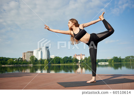 Attractive girl standing in pose king of dance, Asan Natarajasana, at background city landscape with lake and trees 60510614