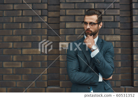 Lost in thoughts. Portrait of handsome bearded businessman in eyeglasses and formal wear thinking about something while standing against brick wall 60531352