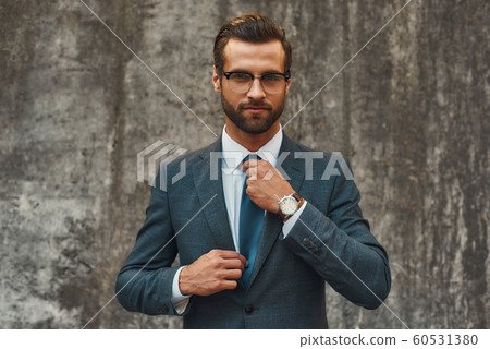 Perfect look. Confident businessman in full suit adjusting his tie and looking at camera while standing against grey stone wall outdoors 60531380